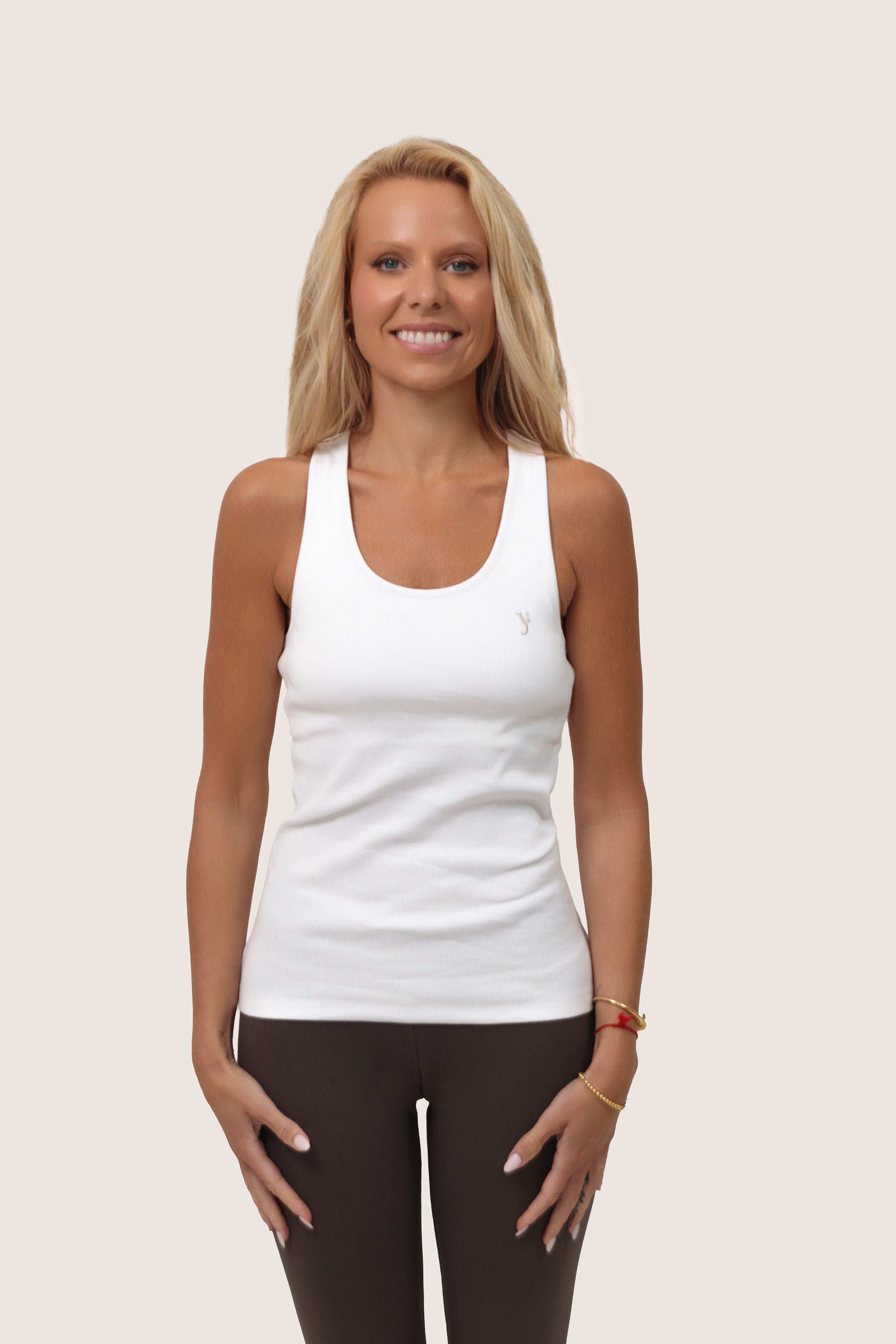 Woman wearing a white tank top from organic cotton and dark leggings on a plain background