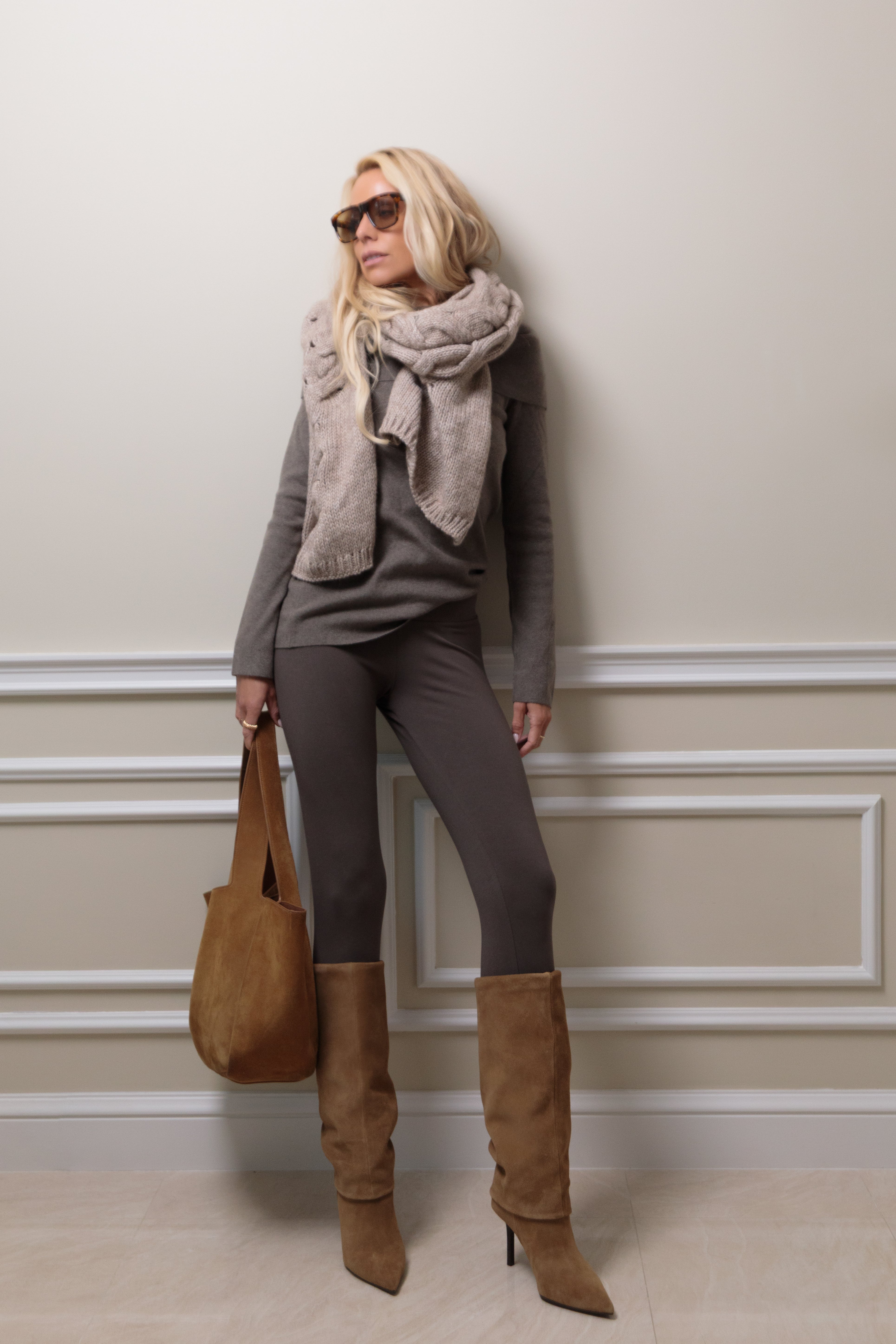 Woman in earthy tone sweater, brown organic cotton leggings, and brown knee-high boots holding a brown bag against a light-colored wall.